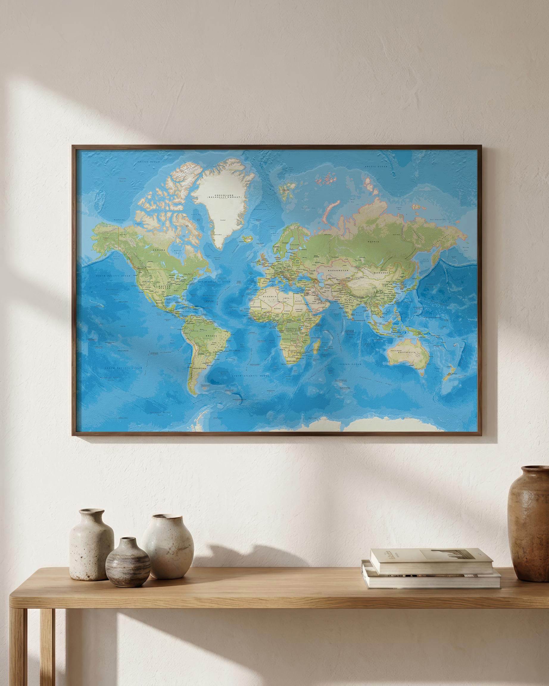 A framed world map art print with blue oceans and green/beige continents, displayed on a white wall above a light wood console table with decorative vases and books.