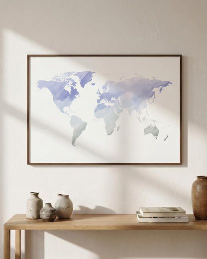 A framed world map art print with a watercolor design in shades of blue, purple, and gray on a white background, hanging on a textured beige wall above a wooden console table with decorative vases and books.