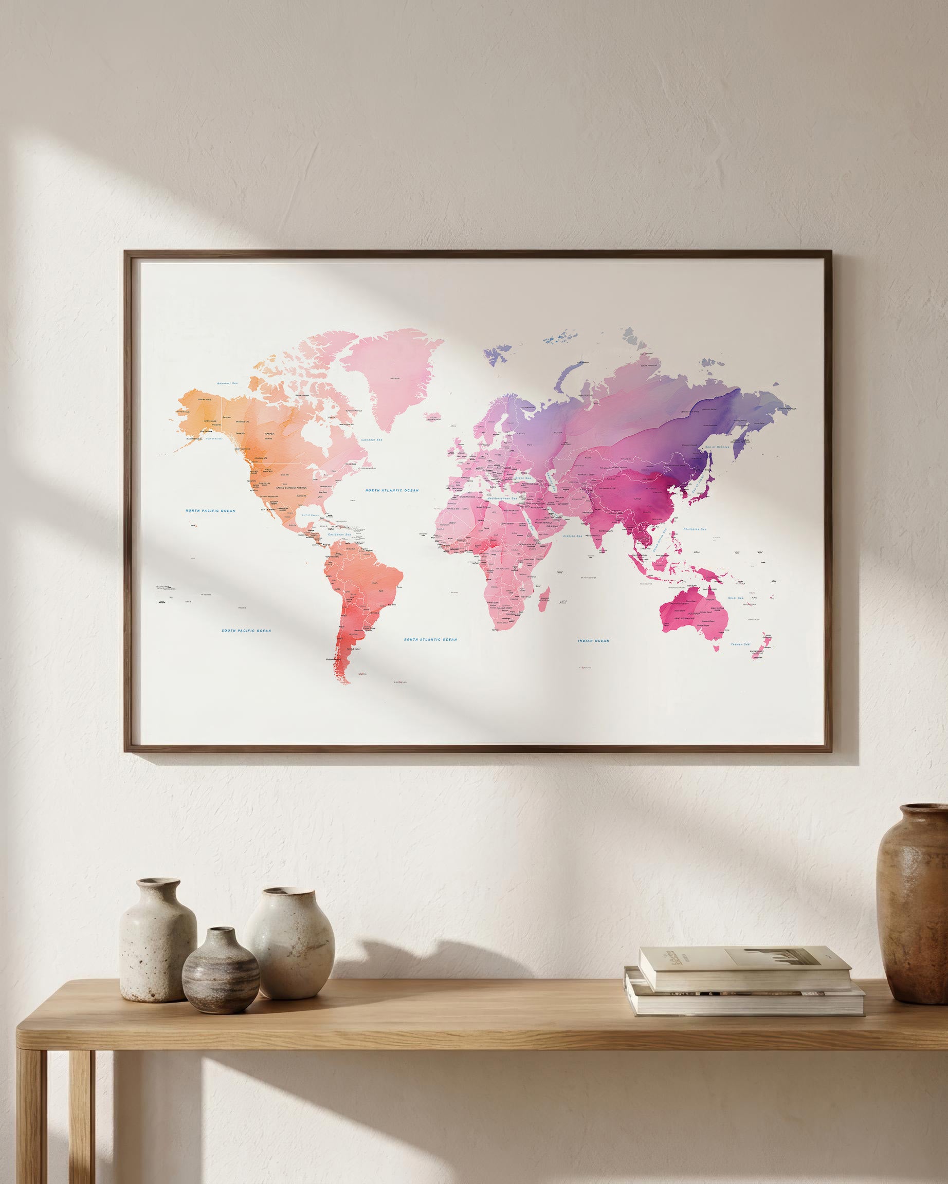 A framed world map art print with a watercolor design in pink, orange, purple, and blue tones, hanging on a light beige wall above a wooden console table with decorative vases and books.