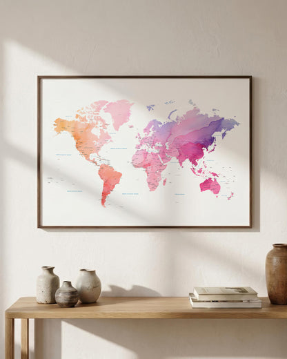 A framed world map art print with a watercolor design in pink, orange, purple, and blue tones, hanging on a light beige wall above a wooden console table with decorative vases and books.