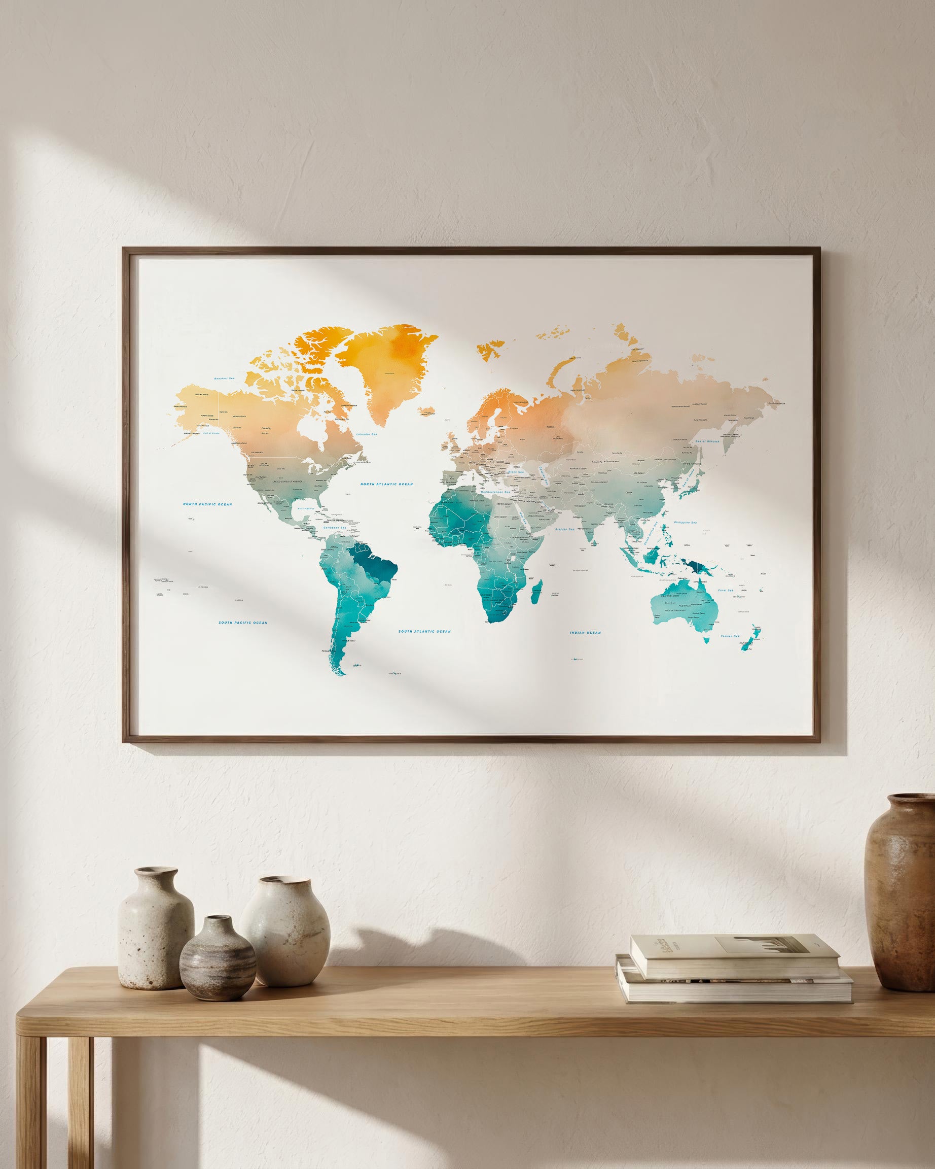 A framed world map art print with a watercolor design in orange, yellow, and teal tones, hanging on a textured white wall above a wooden console table with decorative vases and books.