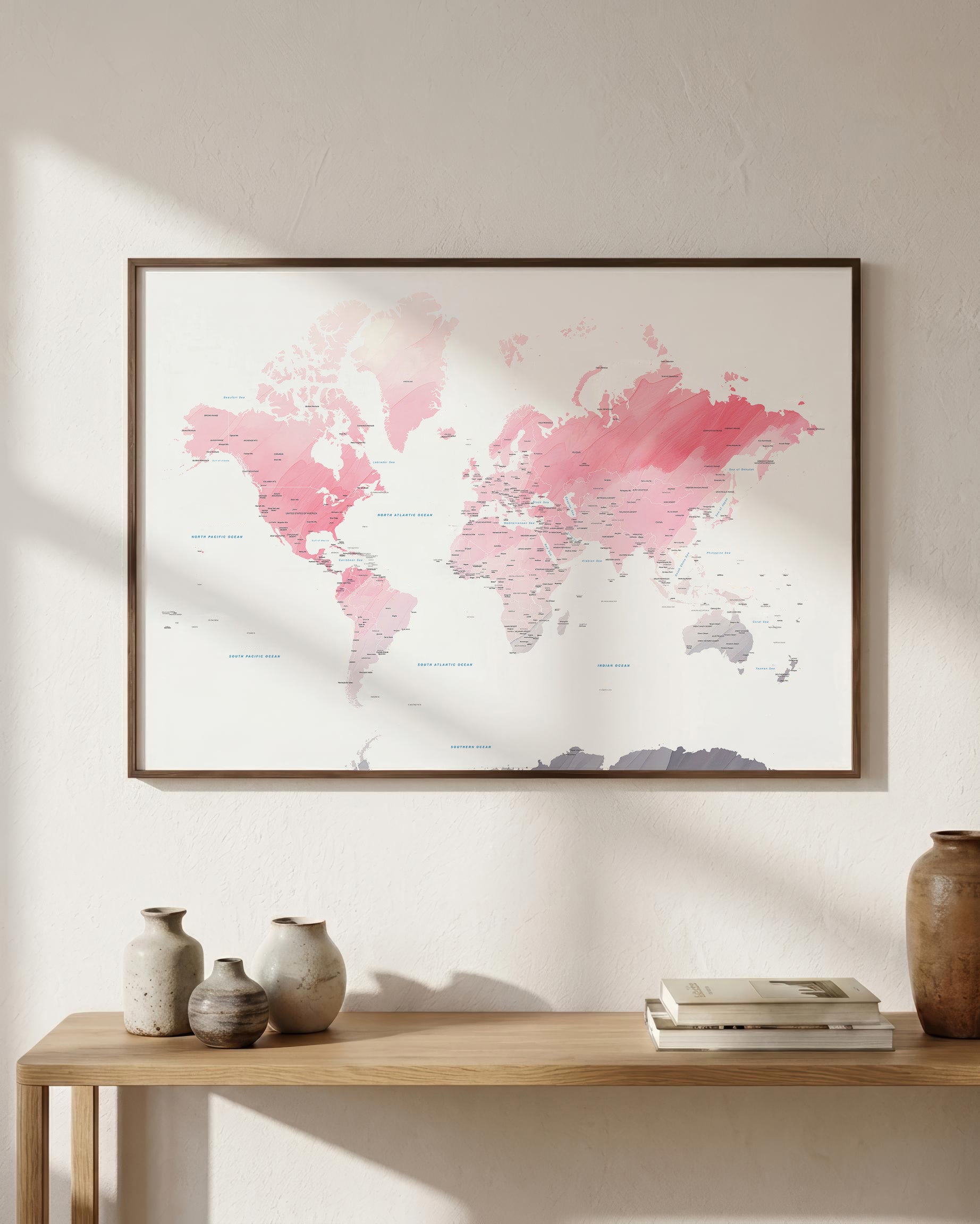 A framed world map art print with pink watercolor continents and a dark wood frame, displayed on a beige wall above a wooden console table with decorative vases and books.
