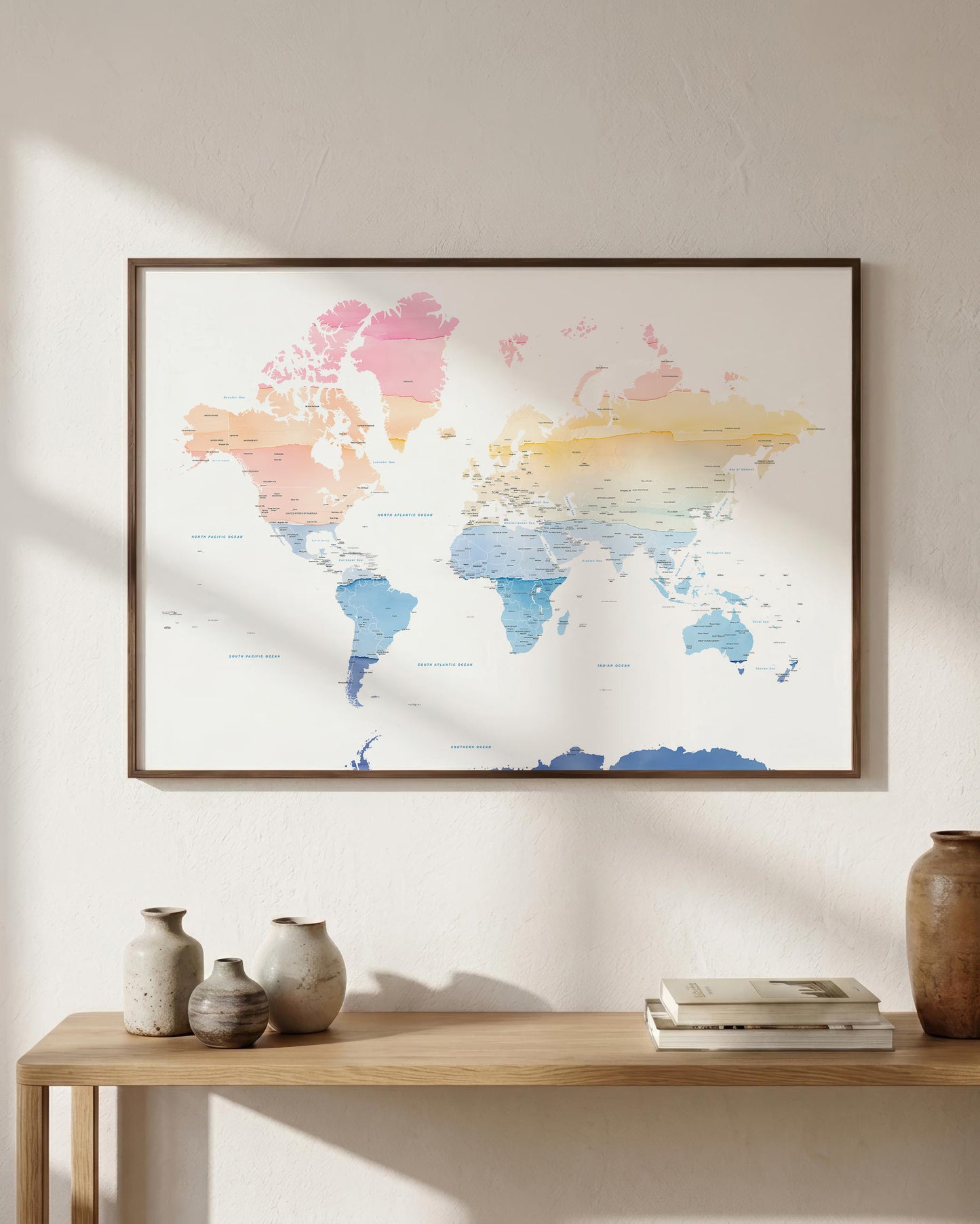 A framed world map art print with pastel watercolor continents in pink, blue, yellow, and peach tones, hanging on a light beige wall above a wooden console table with decorative vases and books.