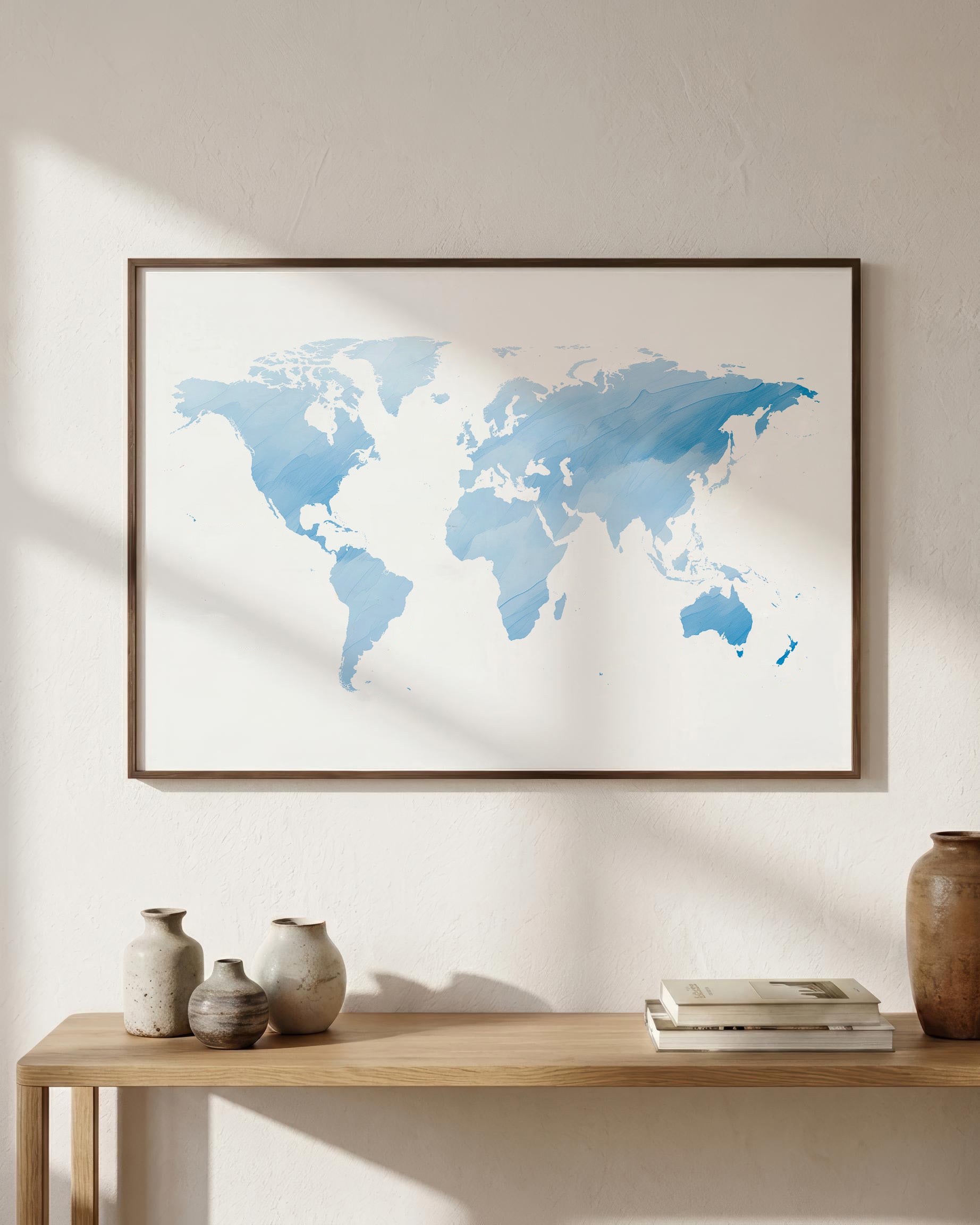 A framed world map art print in shades of blue on a white background, hanging on a beige wall above a wooden console table with decorative vases and books.