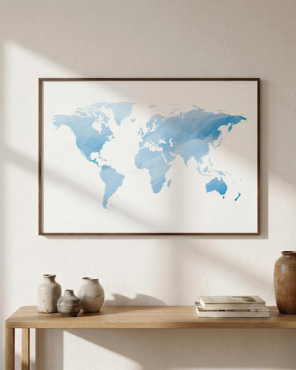 A framed world map art print in shades of blue on a white background, hanging on a beige wall above a wooden console table with decorative vases and books.