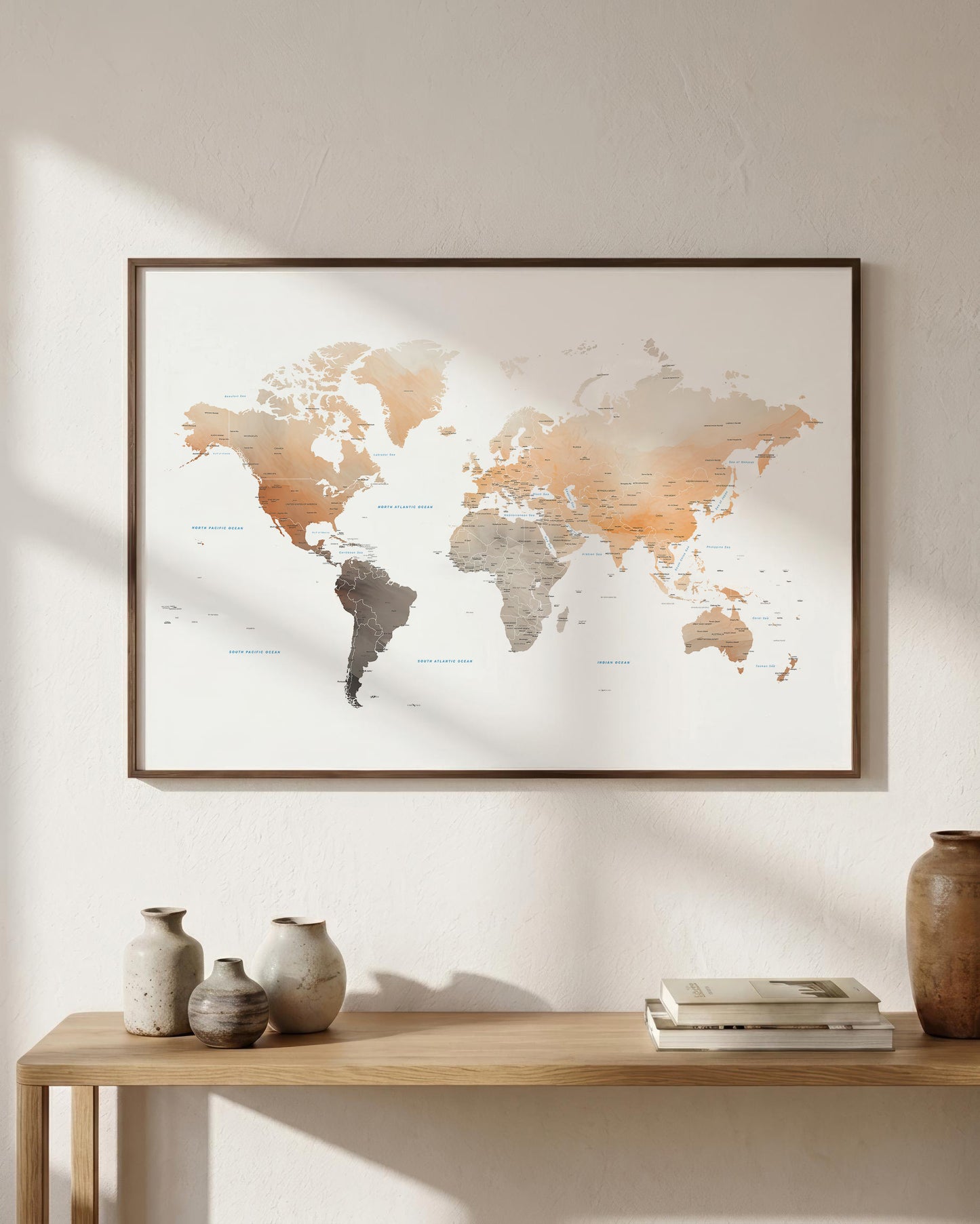 A framed world map print with a minimalist design hangs on a light beige wall above a wooden console table styled with vases and books. The map features watercolor-style continents in warm beige, brown, and gray tones.