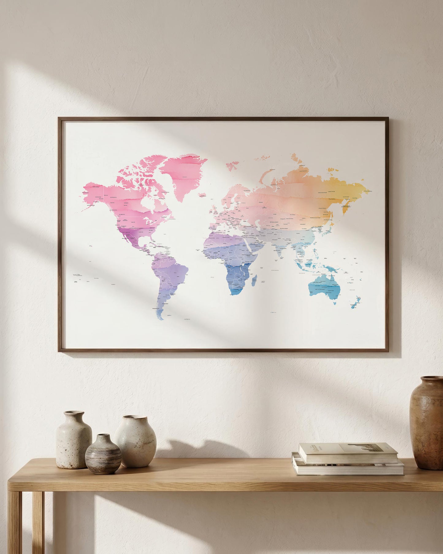 A framed world map art print with a pastel multicolor gradient hangs on a textured off-white wall above a wooden console table styled with vases and books.