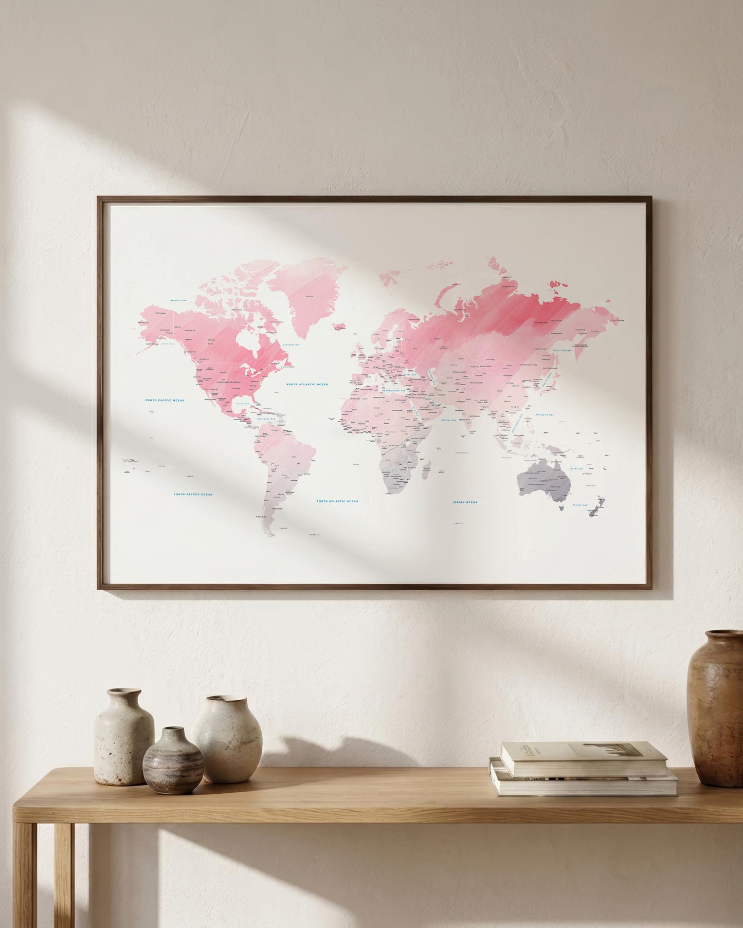 A framed world map print with pink and gray watercolor-style continents hangs on a light beige wall above a wooden console table with vases and books.