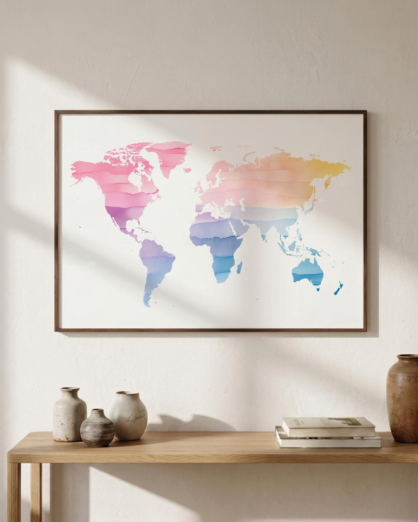 A framed world map art print with a watercolor design in pink, purple, blue, and yellow tones, hanging on a textured beige wall above a wooden console table with decorative vases and books.