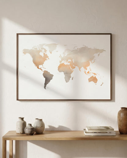A framed world map art print with a minimalist beige, gray, and terracotta watercolor design, hanging on a textured off-white wall above a wooden console table with decorative vases and books.