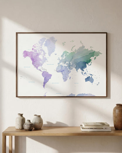 A framed world map art print with a watercolor design in shades of purple, blue, and green, hanging on a textured beige wall above a wooden console table with decorative vases and books.