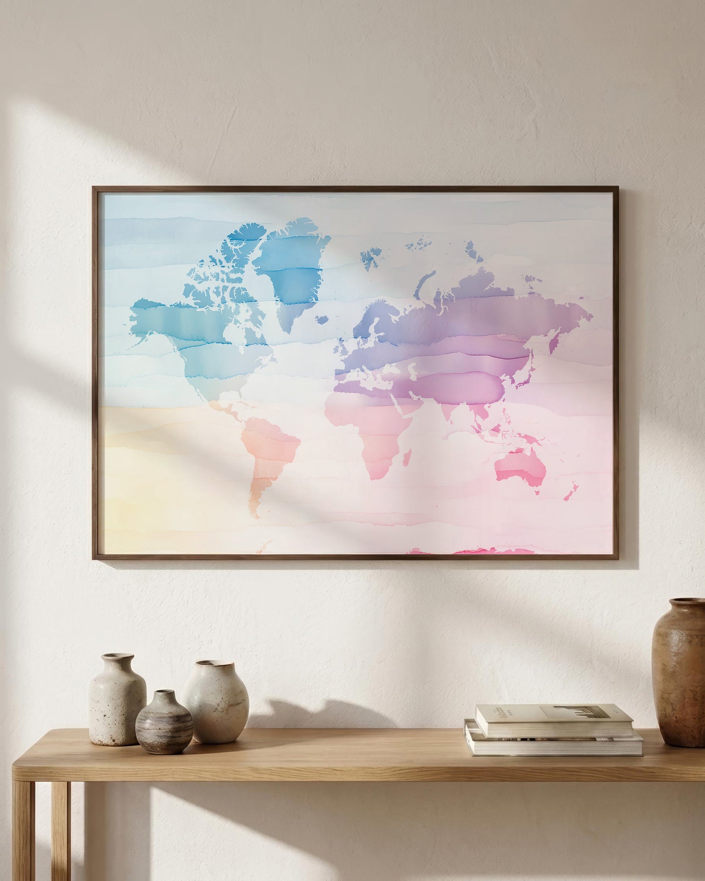 A framed world map art print with a pastel watercolor design in shades of blue, pink, purple, and yellow, hanging on a textured beige wall above a wooden console table with decorative vases and books.