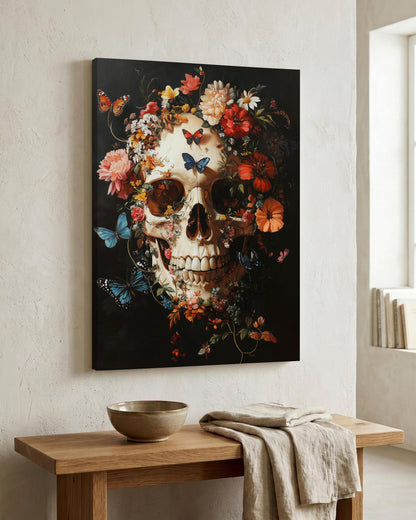 A vertical canvas wall art print featuring a detailed skull surrounded by colorful flowers and butterflies on a black background, displayed on a textured off-white wall above a wooden console table with a bowl and draped cloth.
