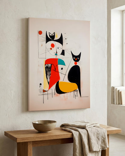 A framed abstract wall art print featuring stylized cats in black, red, yellow, and white on a pale pink background, hanging on a textured off-white wall above a wooden console table with a bowl and draped cloth.