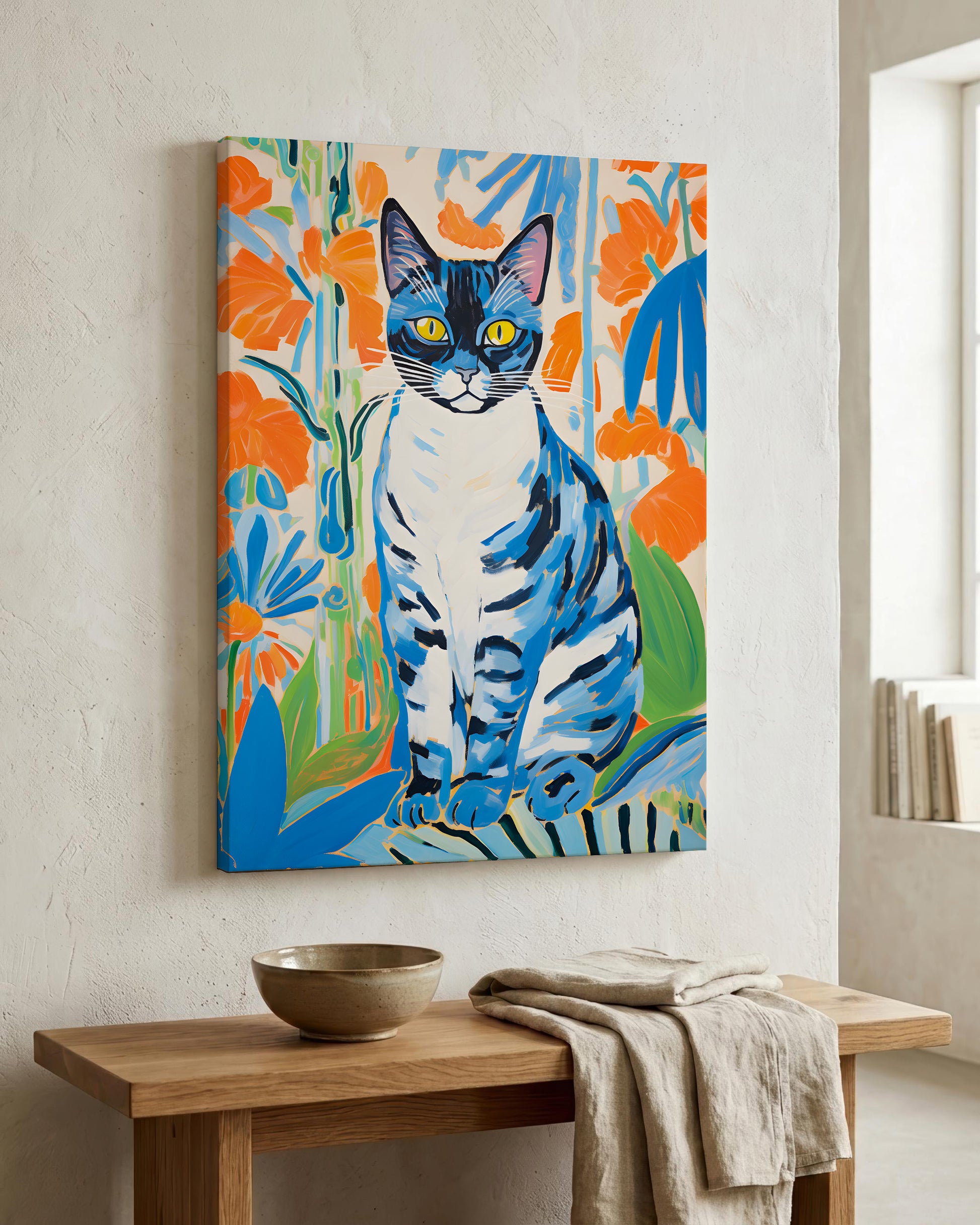 A colorful canvas wall art print depicting a blue and white cat surrounded by bright orange and blue flowers, displayed on a textured off-white wall above a wooden console table with a bowl and draped cloth.