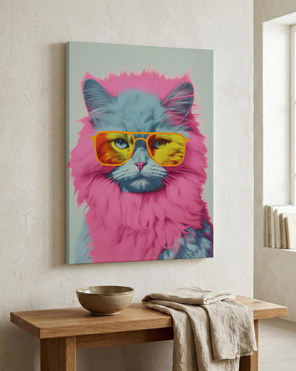 A square wall art print showing a fluffy cat with a bright pink mane and yellow sunglasses, displayed on a textured off-white wall above a wooden console table with a bowl and cloth.