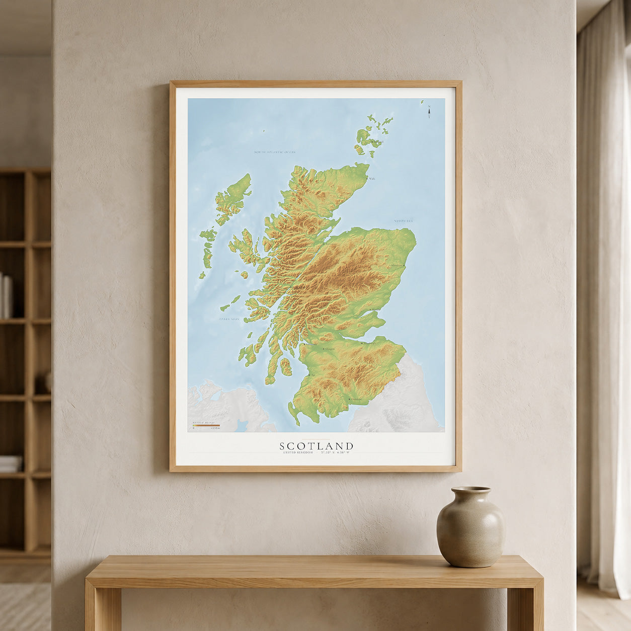 A framed topographic map print of Scotland, featuring green and yellow landmasses against a light blue sea, hanging on a beige wall above a wooden console table with a ceramic vase.