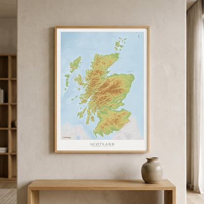 A framed topographic map print of Scotland, featuring green and yellow landmasses against a light blue sea, hanging on a beige wall above a wooden console table with a ceramic vase.