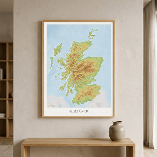 A framed topographic map print of Scotland, featuring green and yellow landmasses against a light blue sea, hanging on a beige wall above a wooden console table with a ceramic vase.