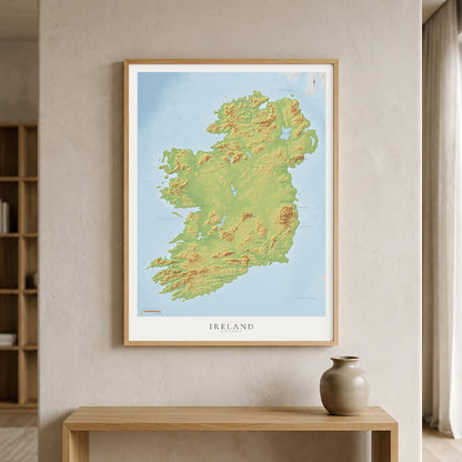 A framed topographical map of Ireland with green and yellow landmasses and a light blue sea, displayed on a beige wall above a wooden console table with a small vase.