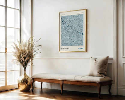 A framed art print of a light blue and gray Berlin street map hangs on a white wall above a wooden bench with a beige cushion in a bright, modern room.