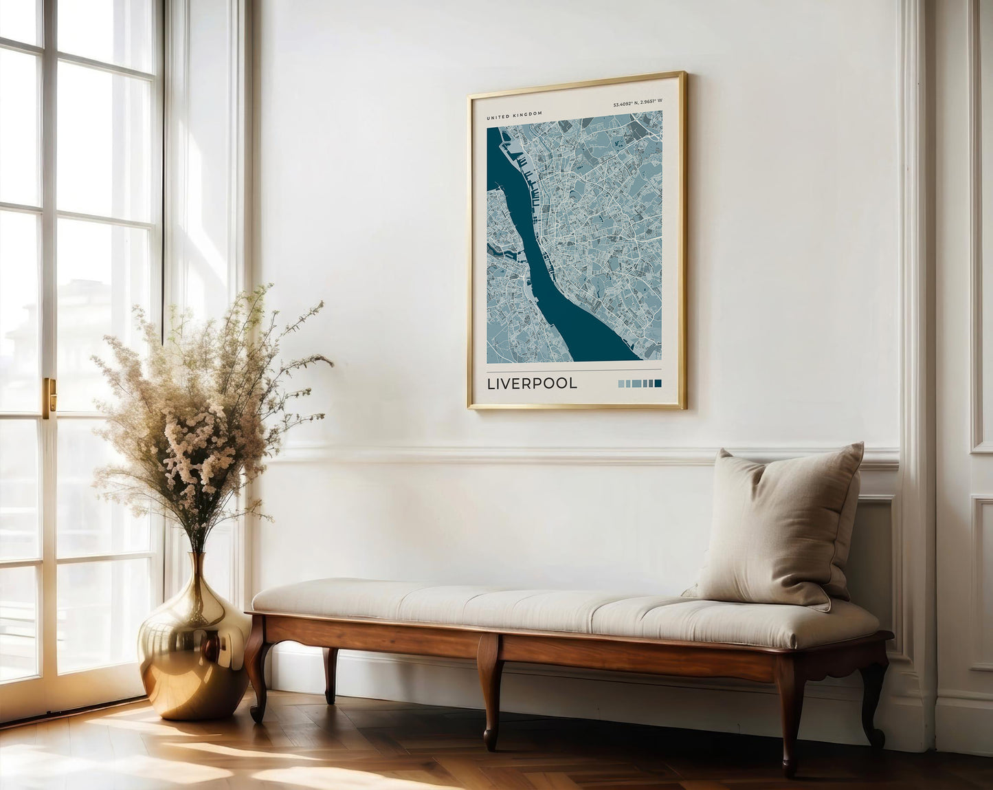A framed Liverpool city map print hangs on a white wall above a wooden bench with a cream cushion in a bright, sunlit room. A tall metallic vase with dried stems stands near the window.