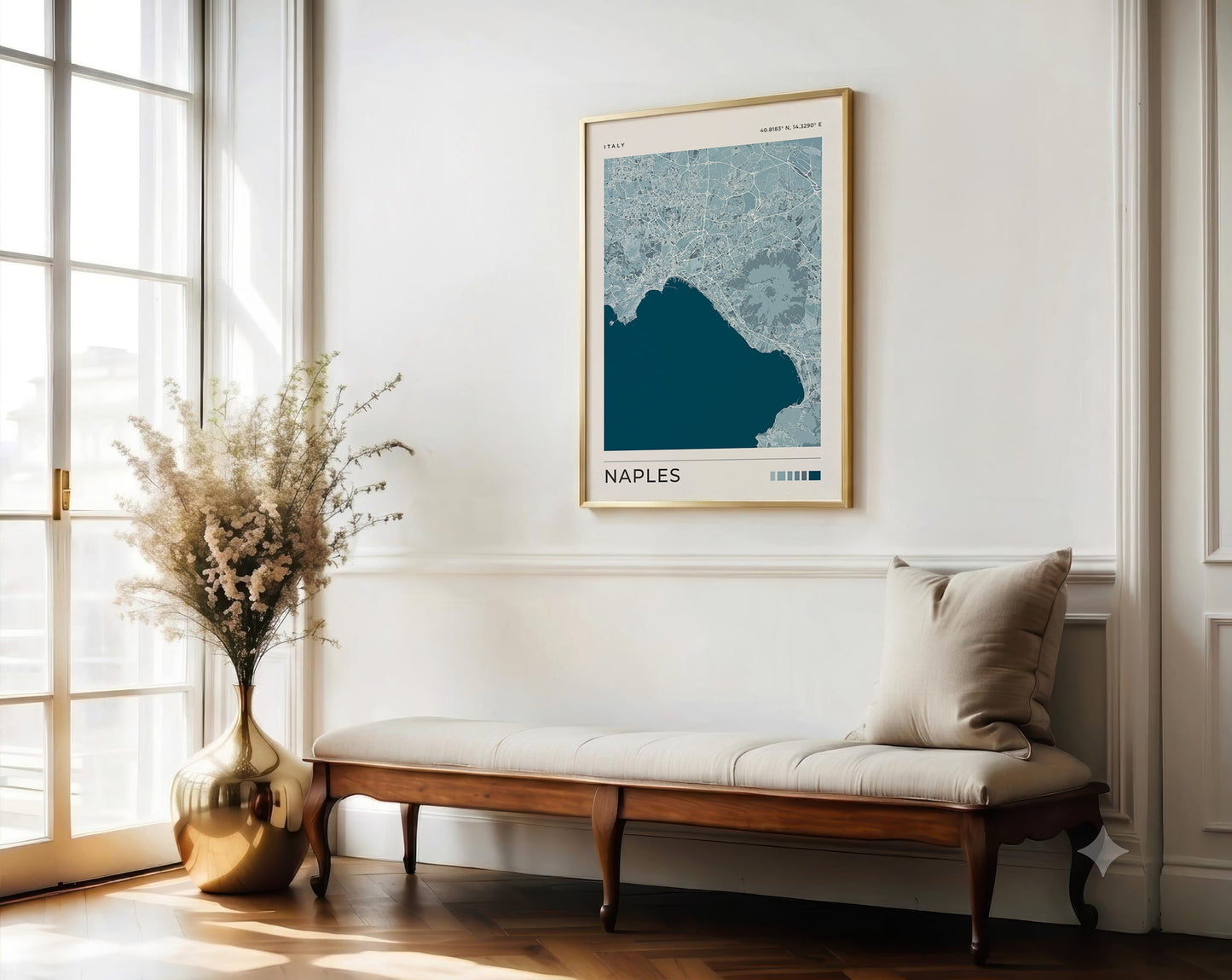 A framed Naples map art print hangs on a white wall above a wooden bench with a beige cushion in a bright, sunlit room. A large gold vase with dried flowers sits beside the window.