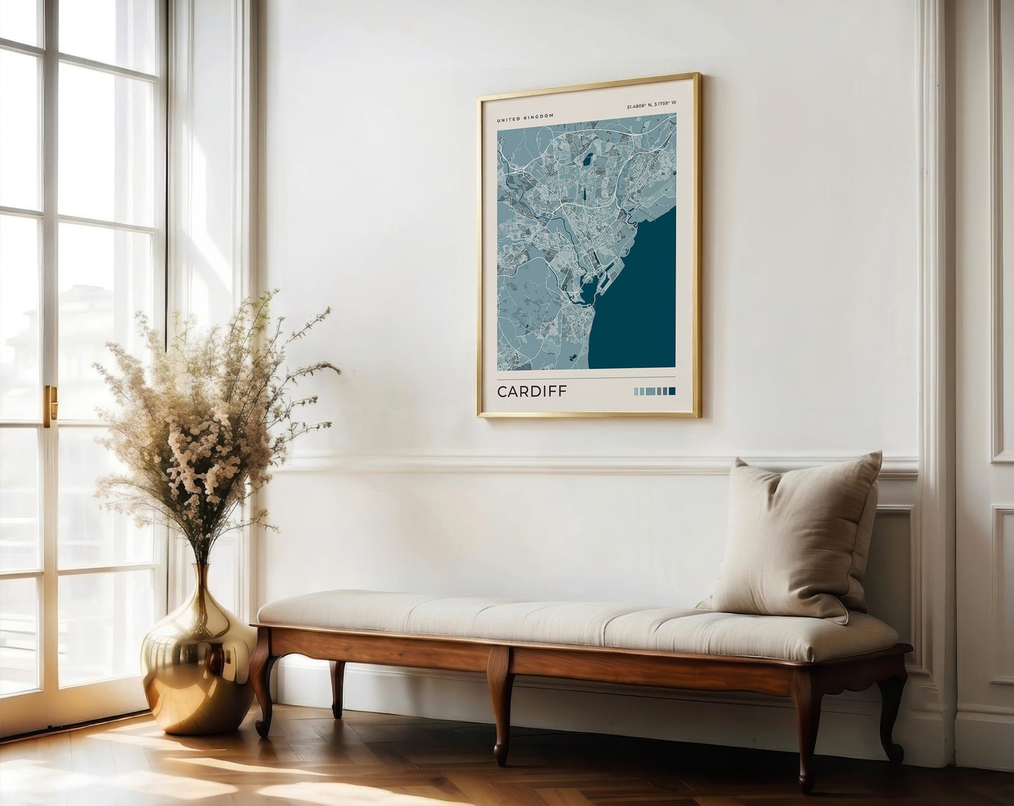 A framed map print of Cardiff, Wales, hanging on a white wall in a bright, modern room with a wooden bench and a large vase of dried flowers.