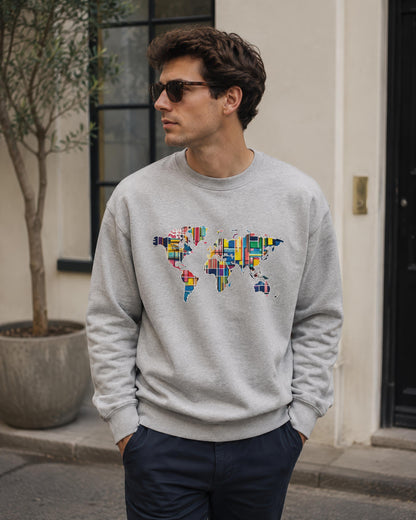 A man wearing a light gray crewneck sweatshirt with a colorful world map graphic made of various national flags, paired with dark navy pants and sunglasses, standing outdoors in front of a building.