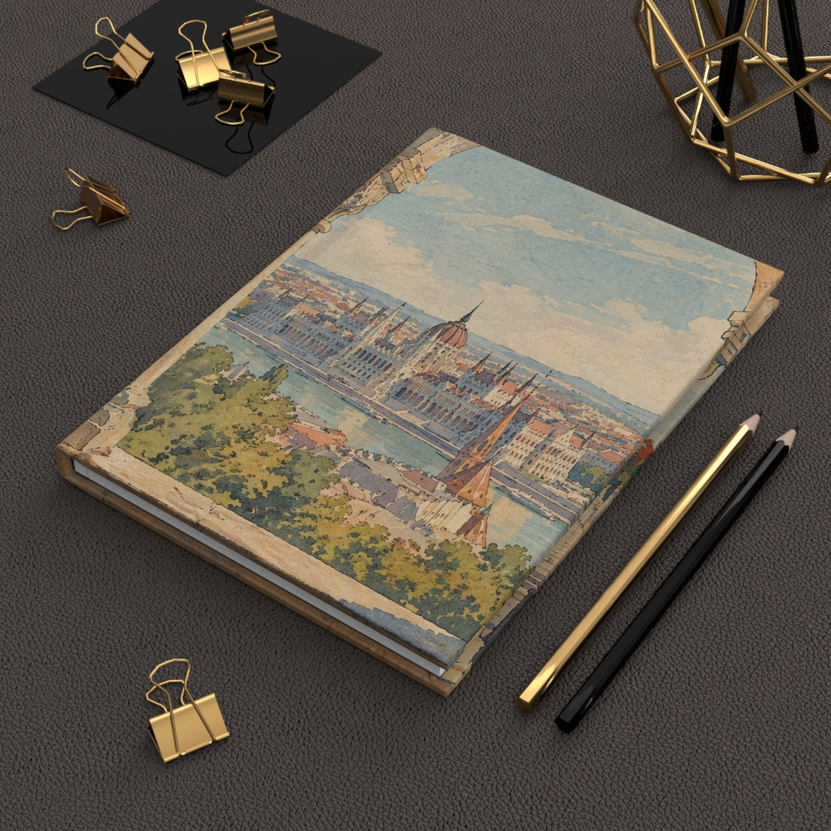 A hardcover notebook featuring a vintage-style illustration of Budapest's cityscape on the cover, styled on a dark textured surface with gold binder clips, pencils, and a geometric gold decor piece.