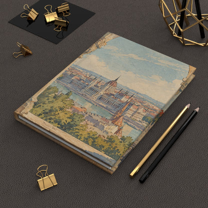 A hardcover notebook featuring a vintage-style illustration of Budapest's cityscape on the cover, styled on a dark textured surface with gold binder clips, pencils, and a geometric gold decor piece.