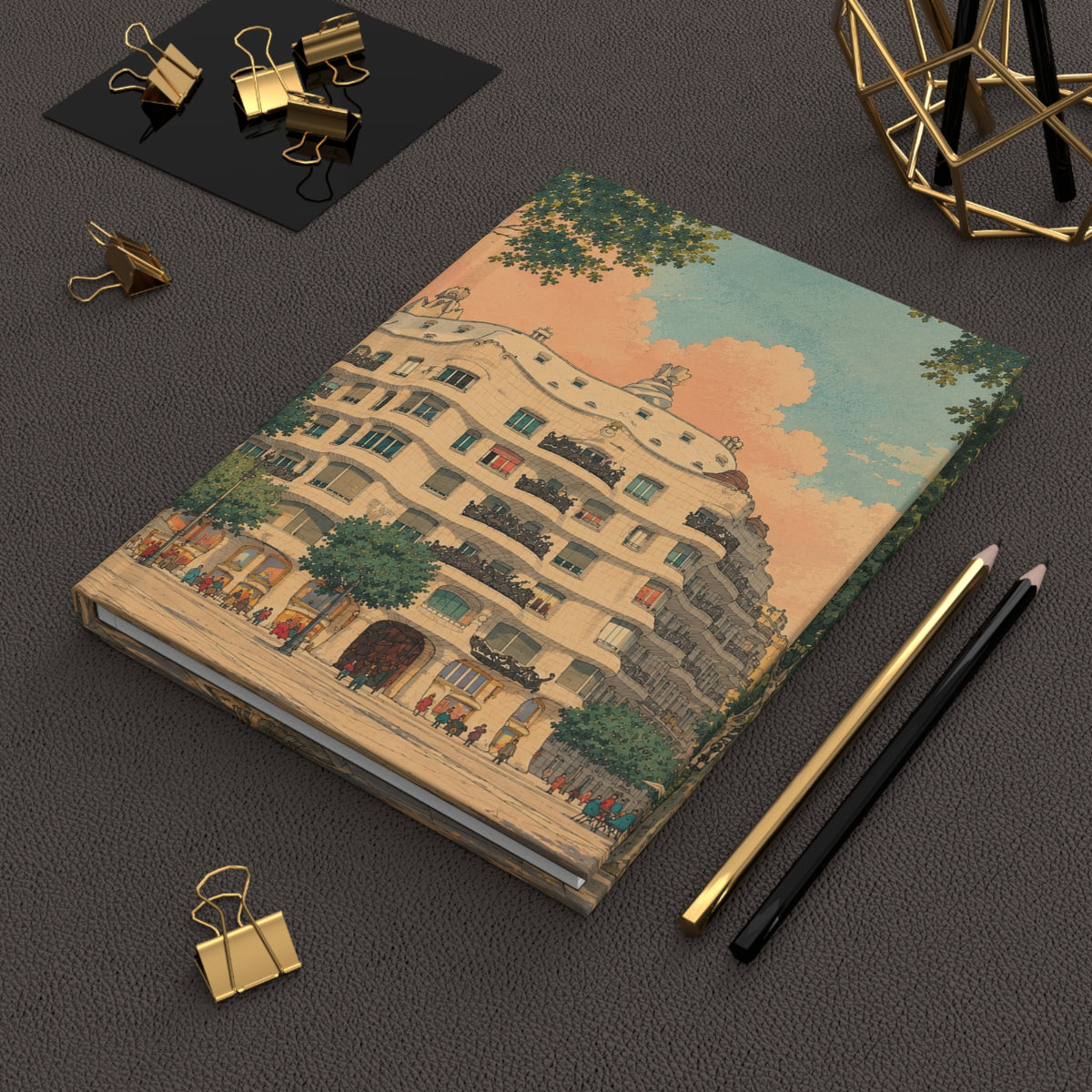A notebook with a vintage-style architectural illustration on the cover, styled on a dark textured surface with gold binder clips, pencils, and a geometric gold decor piece.