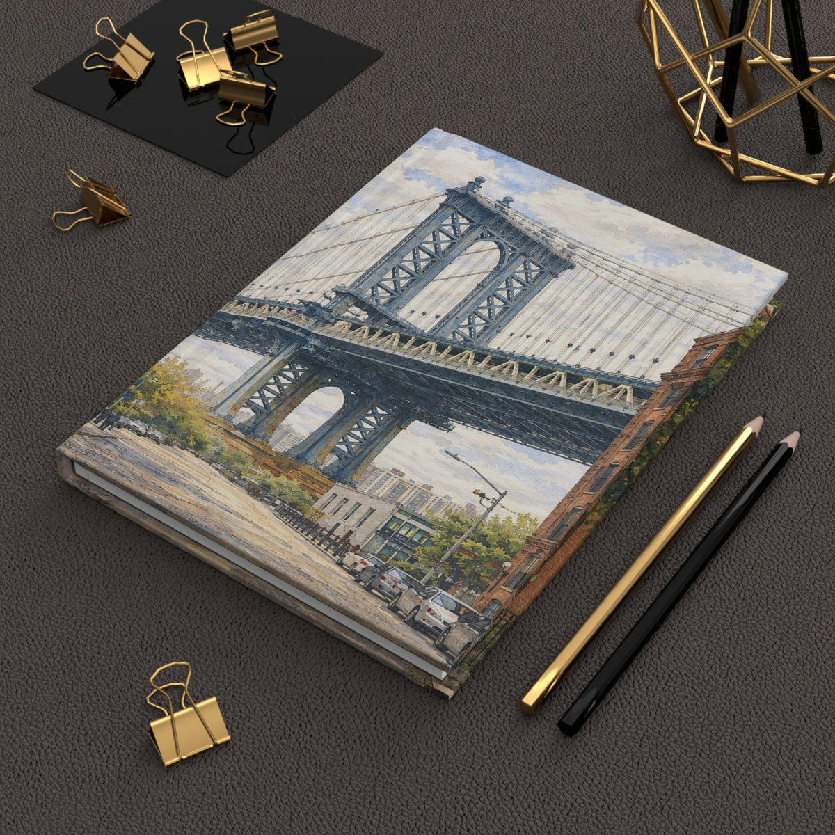 A hardcover notebook featuring a watercolor-style illustration of the Manhattan Bridge in New York City, styled on a dark textured surface with gold and black office accessories.