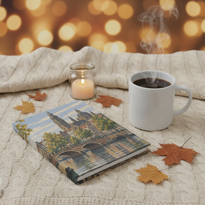 A cozy lifestyle scene featuring a hardcover notebook with a Parisian bridge and cathedral illustration, styled on a cream cable-knit blanket with autumn leaves, a lit candle in a glass jar, and a white mug of coffee with steam rising against a warm golden bokeh background.
