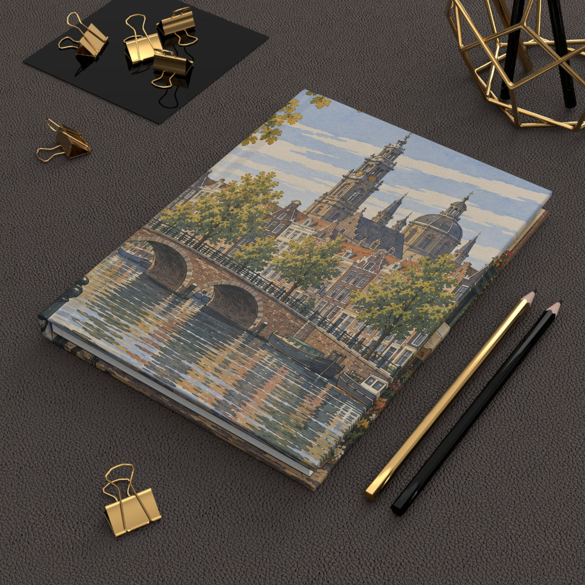 A hardcover notebook featuring a detailed painting of Amsterdam canals and historic buildings, styled on a dark textured surface with gold and black office accessories.