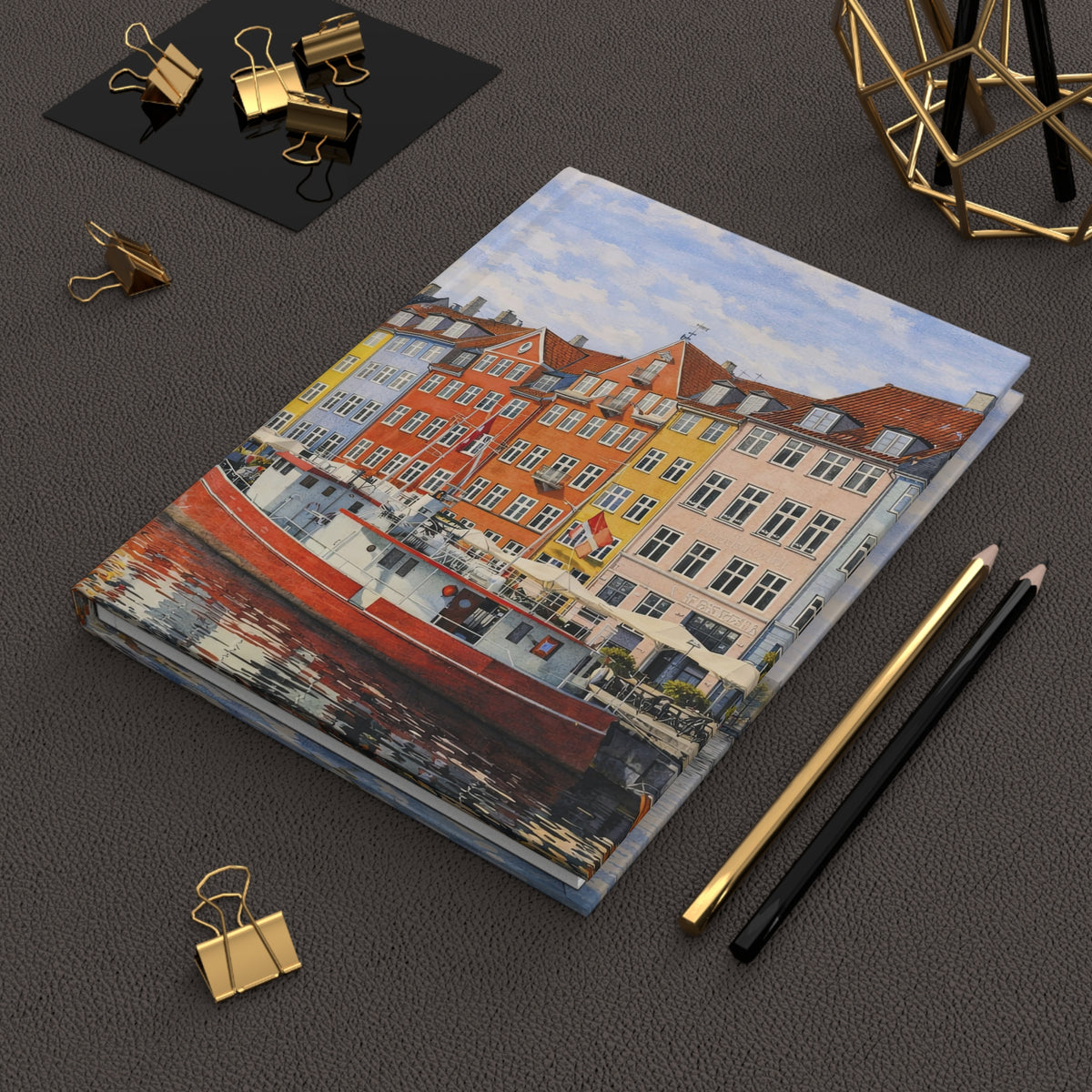 A notebook featuring a colorful watercolor-style painting of a canal with red-roofed buildings and a red boat, styled on a dark textured surface with gold binder clips, pencils, and a geometric gold decor object.