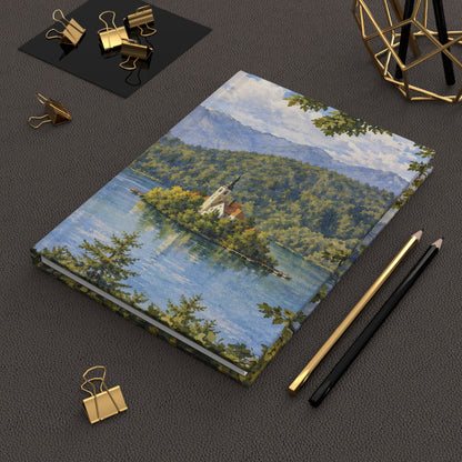 A hardcover notebook with a scenic landscape illustration of a lake, forest, and mountains, styled on a dark textured surface with gold binder clips, pencils, and a geometric gold decor piece.