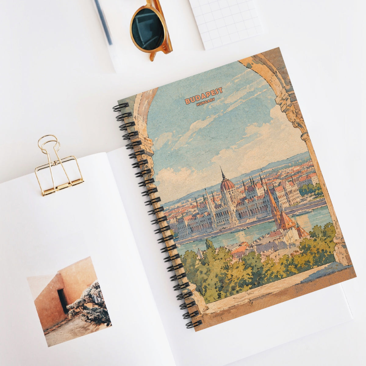 A spiral-bound notebook with a vintage-style Budapest cityscape cover design, styled on a white surface with an open page, sunglasses, a gold binder clip, and a pen nearby.