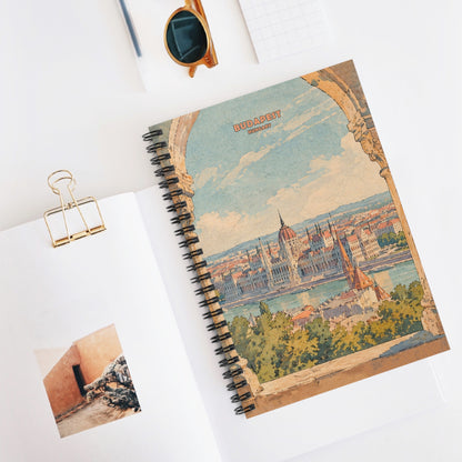 A spiral-bound notebook with a vintage-style Budapest cityscape cover design, styled on a white surface with an open page, sunglasses, a gold binder clip, and a pen nearby.