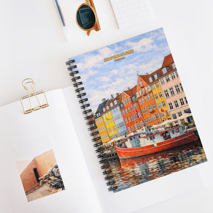 A spiral-bound notebook with a colorful cover illustration of Nyhavn, Copenhagen, shown in a styled flat-lay on a white desk next to sunglasses, a gold binder clip, and another open notebook.
