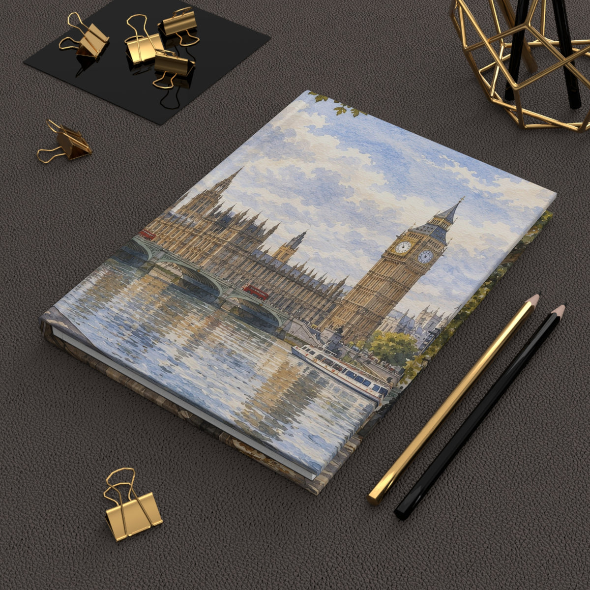 A notebook with a printed illustration of Big Ben and the Houses of Parliament across the River Thames, styled on a dark textured surface with gold and black pencils, binder clips, and a geometric gold decor object.