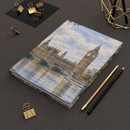 A notebook with a printed illustration of Big Ben and the Houses of Parliament across the River Thames, styled on a dark textured surface with gold and black pencils, binder clips, and a geometric gold decor object.