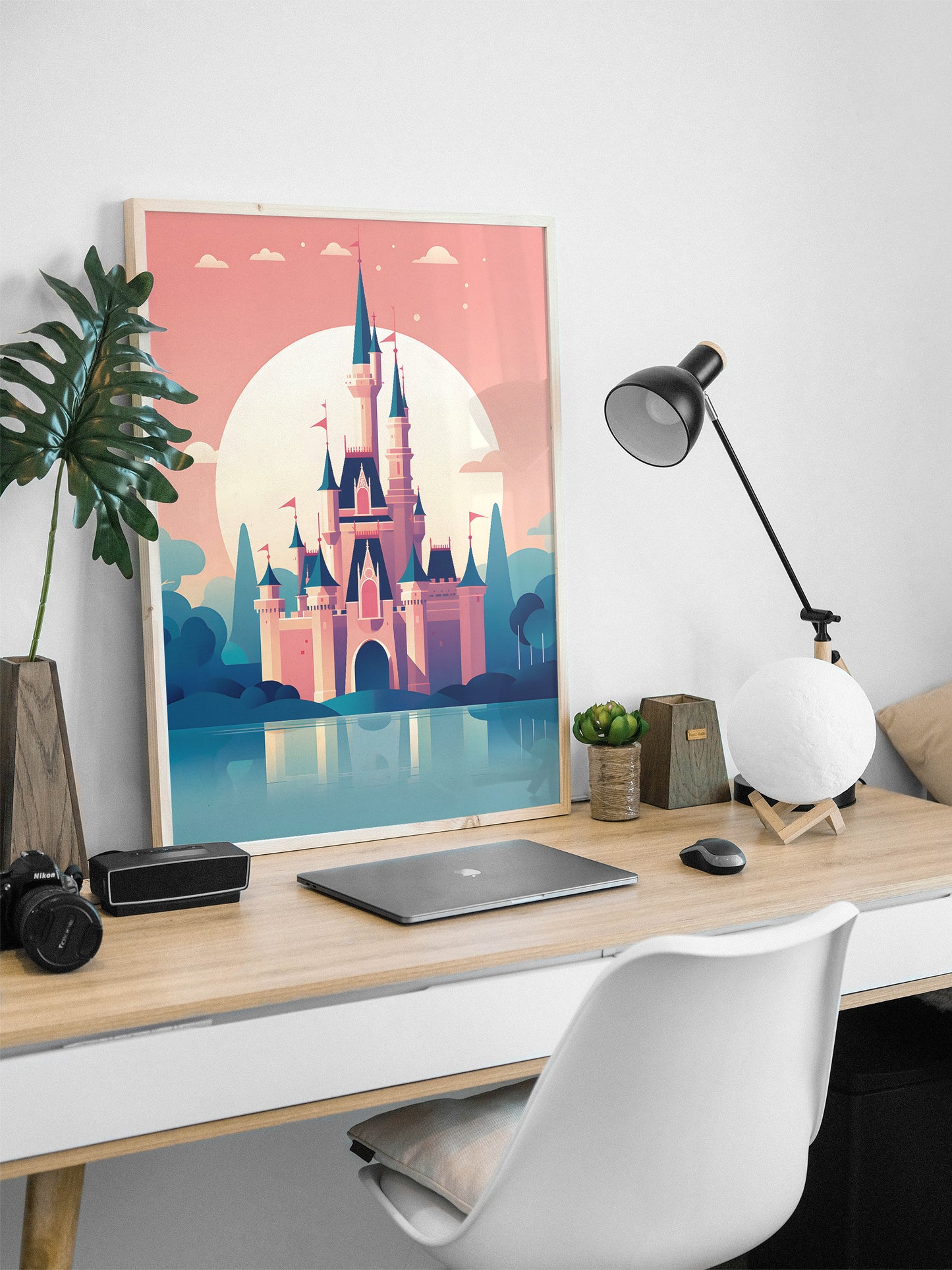 A framed art print of a whimsical castle in a pink sunset landscape, displayed on a light wood desk in a modern home office setting with a laptop, lamp, and chair.