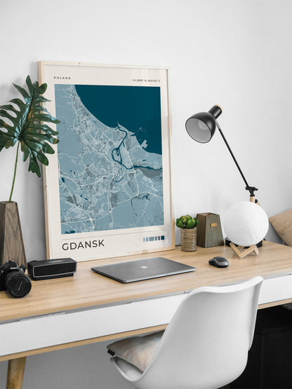 A framed art print of a Gdańsk city map in shades of blue and gray, styled on a light wood desk with a laptop, lamp, and plant.