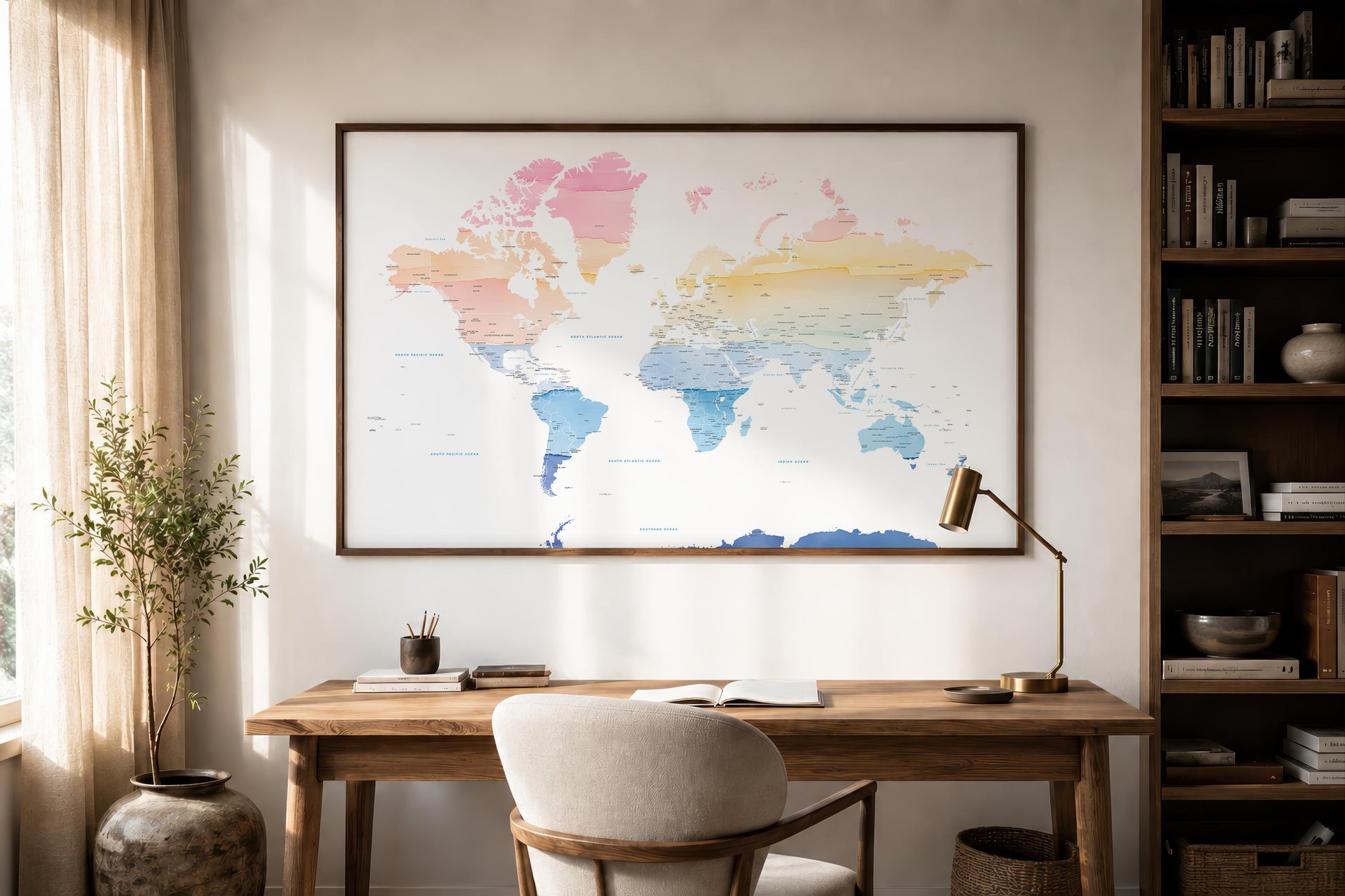 A framed world map poster with pastel watercolor colors hangs on a light beige wall above a wooden desk in a styled home office setting with a chair, lamp, and bookshelf.