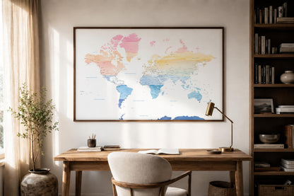 A framed world map poster with pastel watercolor colors hangs on a light beige wall above a wooden desk in a styled home office setting with a chair, lamp, and bookshelf.