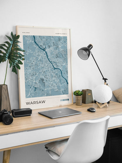 A framed poster of a blue and white map of Warsaw, Poland, displayed on a light wood desk in a modern home office setting with a white chair, lamp, and laptop.