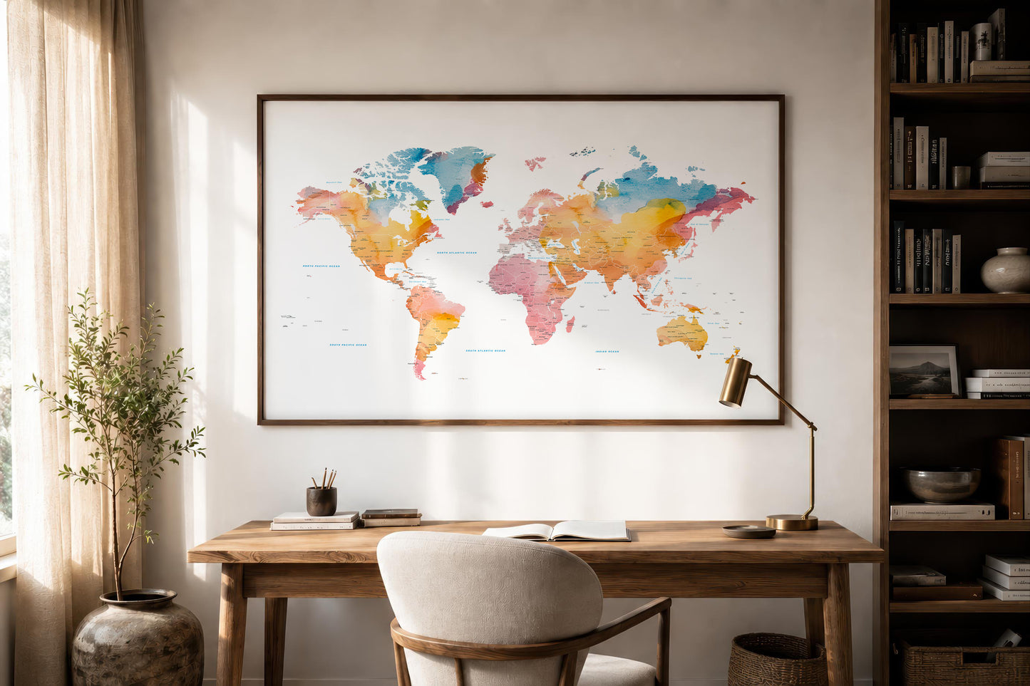 A large framed world map print with colorful watercolor-style continents hangs on a beige wall above a wooden desk in a styled home office setting with a chair, lamp, bookshelf, and plant.