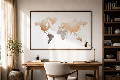 A large framed world map print with a watercolor-style design hangs on a wall above a wooden desk in a styled home office setting with a chair, lamp, and bookshelves.