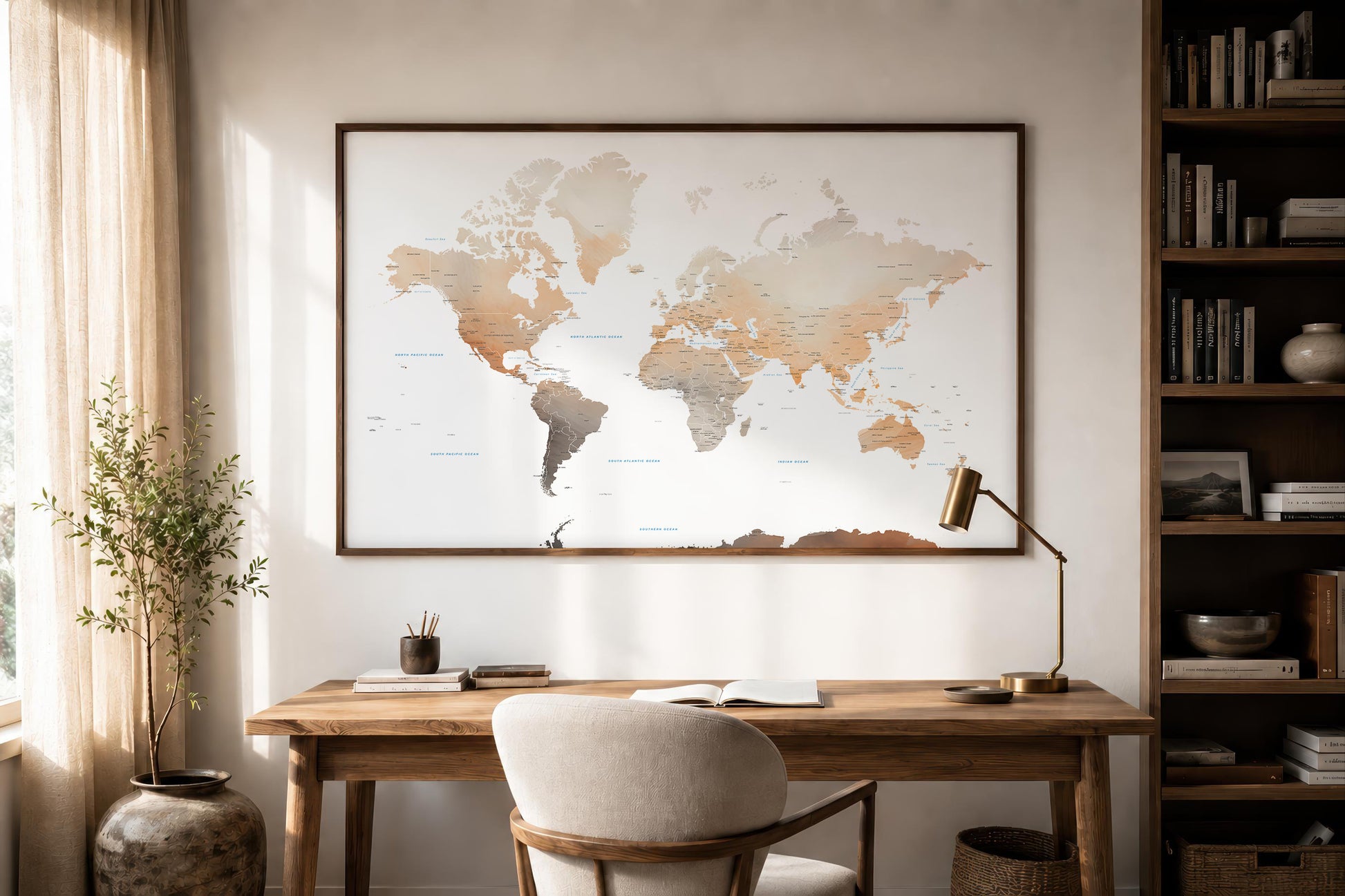 A large framed world map print hangs on a light beige wall above a wooden desk in a styled home office. The room includes a white upholstered chair, a brass desk lamp, a potted plant, and a tall bookshelf filled with books.